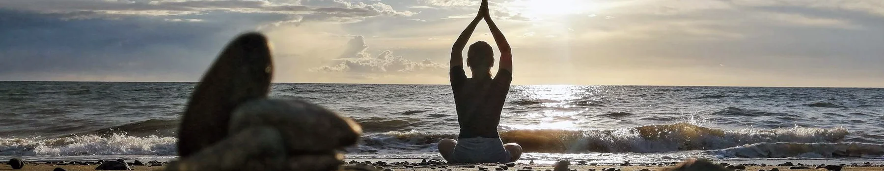 Woman sitting in meditation with her arms raised on the beach at sunrise looking at the water